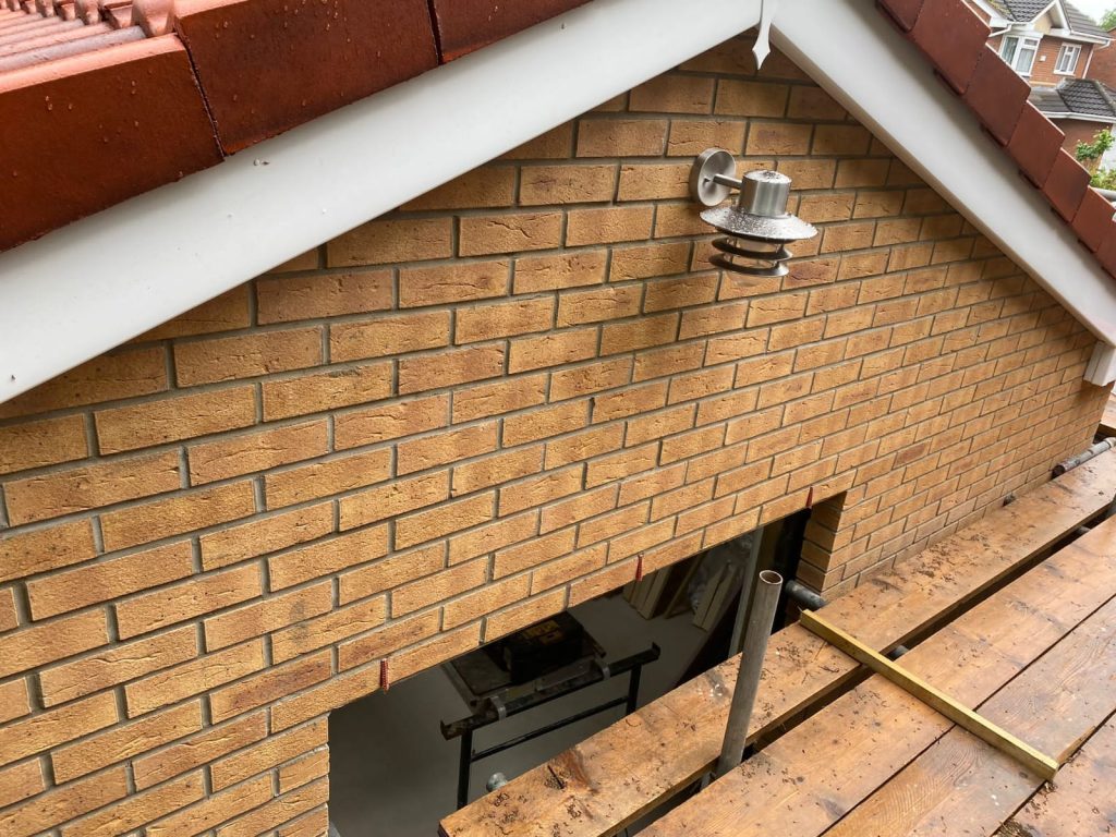 A brick wall under a red-tiled roof features an outdoor wall light. Below is a wooden scaffolding platform. The image is taken from above, showing part of a ladder and materials inside the opening.