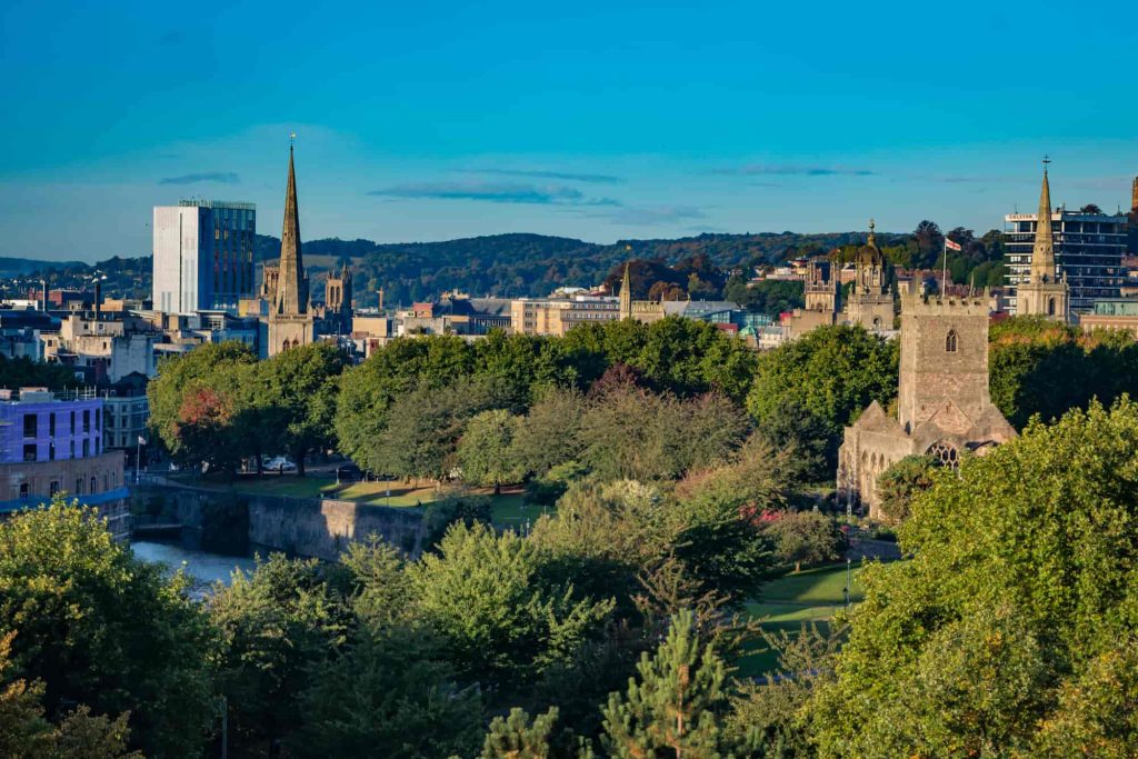 A cityscape view of Bristol, England, featuring green trees, historic churches with tall spires, modern buildings, and distant hills under a clear blue sky.