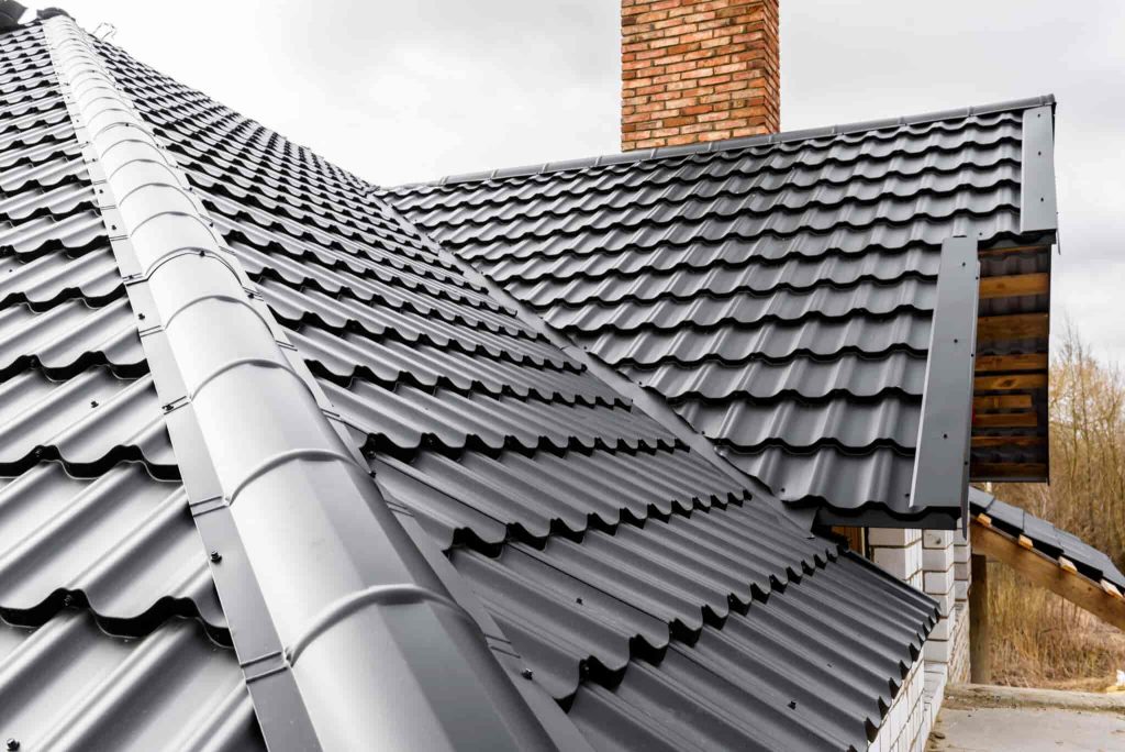 A close-up view of a house roof with dark gray metal tiles and a red brick chimney, set against a cloudy sky. The roofing has a modern, wavy pattern and appears clean and newly installed.