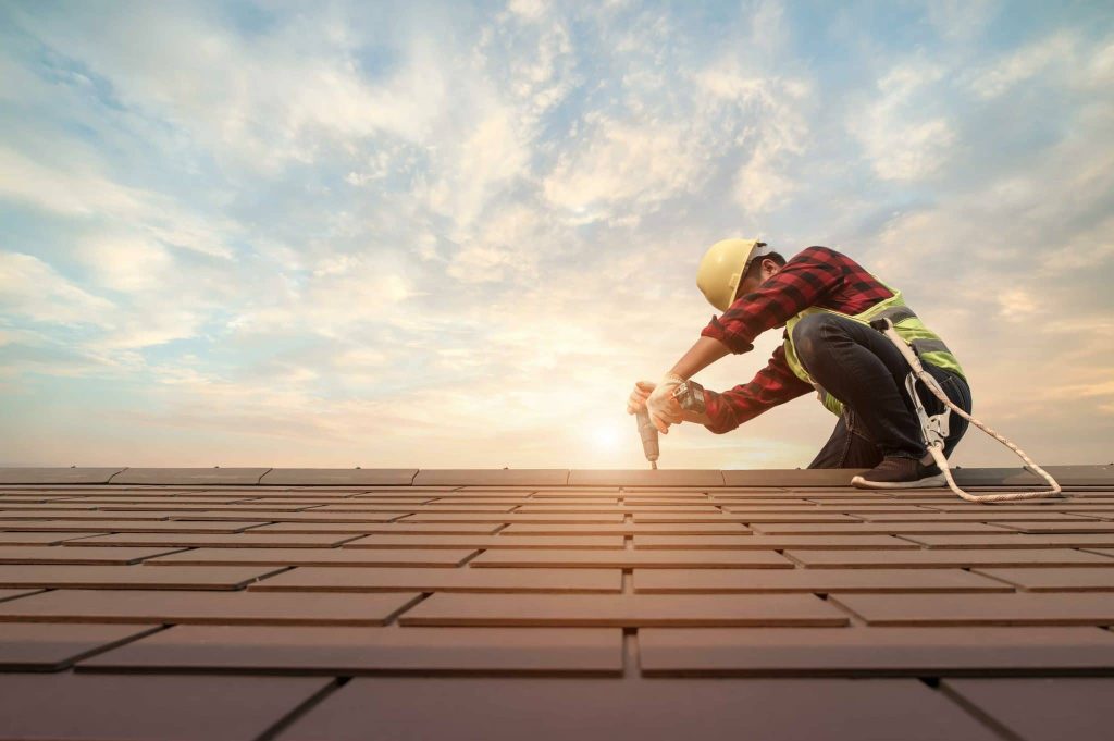 A construction worker wearing a hard hat and safety harness kneels on a roof, using a power drill to secure tiles, with a bright sky and setting sun in the background.
